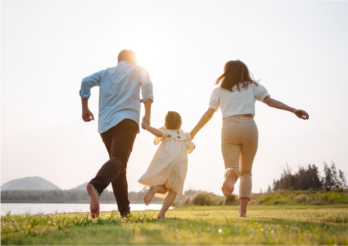 A joyful family of three, holding hands and running barefoot on a grassy field during a sunny day.