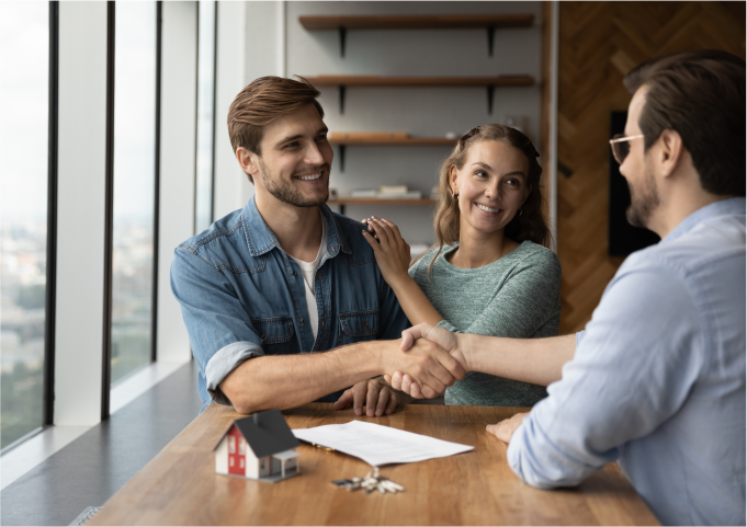 A young couple happily signing a document and shaking hands with a real estate agent in a bright, modern office.